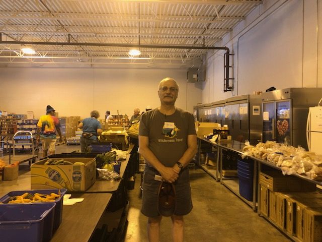 Michael standing in front of aisles of food wearing a shirt and t-shirt at the Jewish Community Pantry.