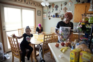 Two children sit at a kitchen table, while an older sibling slices apples at a counter. 