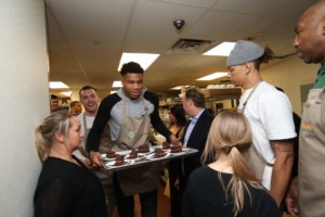  Giannis Antetokounmpo serves dessert, while a large group of people surrounds him. at the 2019 Coach Bud's Assists for Hunger Dinner. 