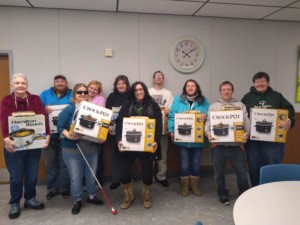 10 individuals holding 10 new crock pots still in the boxes in a classroom setting. 