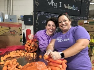 Two women smiling wearing purple shirts and sorting carrots in front of a black board that says 