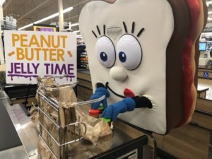 A peanut butter and jelly mascot bagging jars of peanut butter and the grocery store.