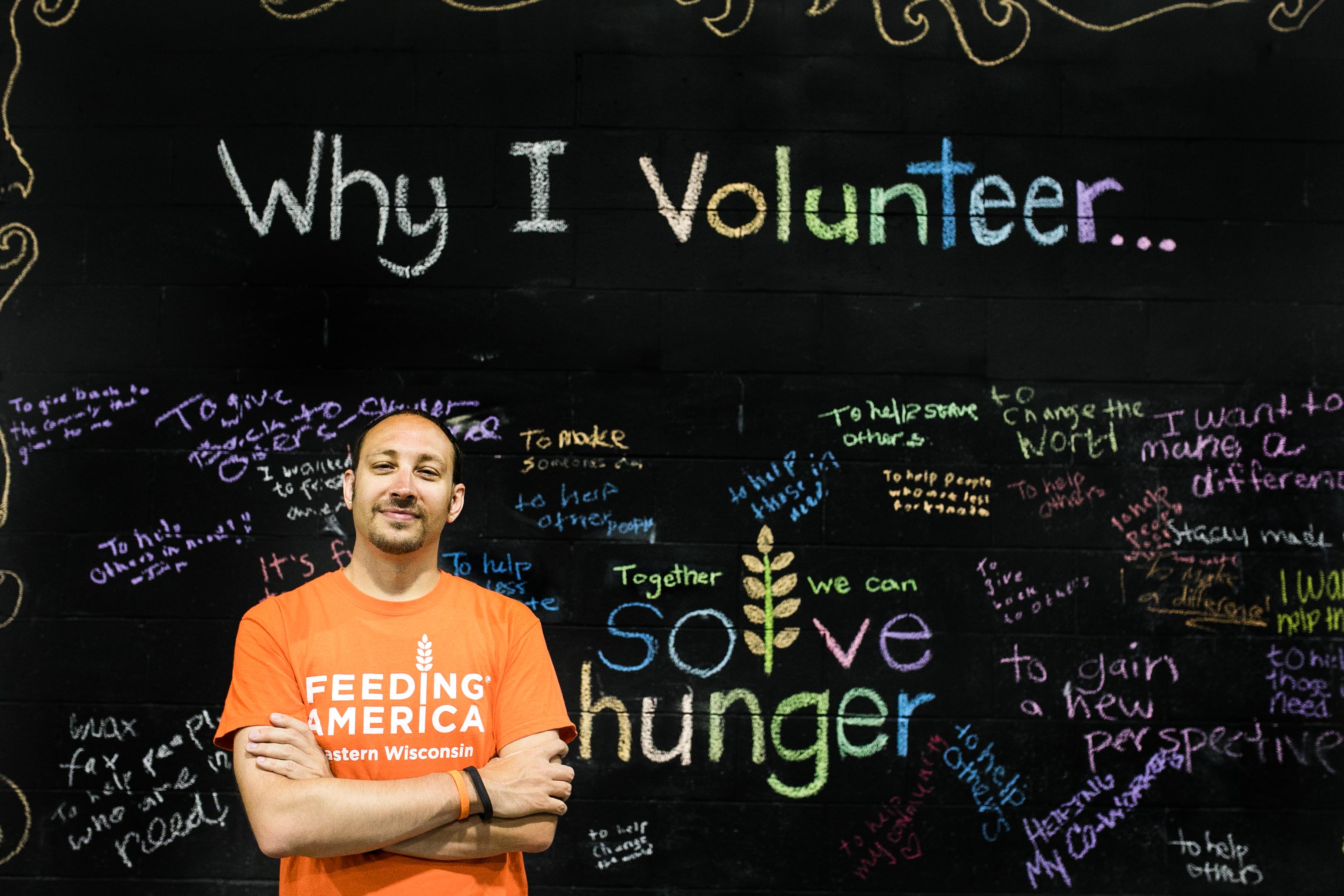 Warren wearing an orange Feeding America Eastern Wisconsin shirt in front of a black chalk board listing reasons why people volunteer. 
