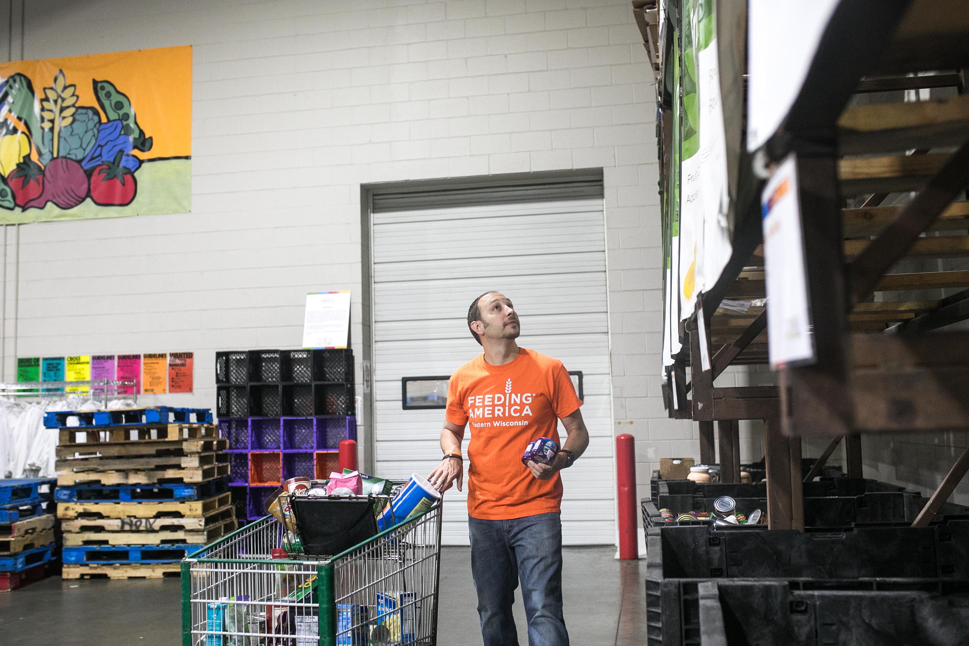 Warren wearing an orange Feeding America Eastern Wisconsin shirt while pushing a cart full of food to sort into bins.