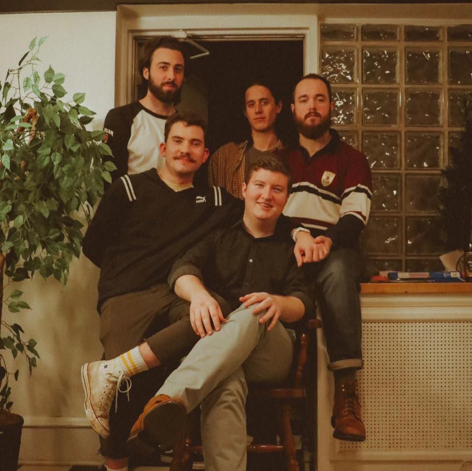 The five members of the band Motel Breakfast stand in a doorway with a plant to their right. 