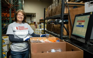 A woman wearing a Team Rubicon "greyshirt" poses by a box of donations, ready to scan them into the system.