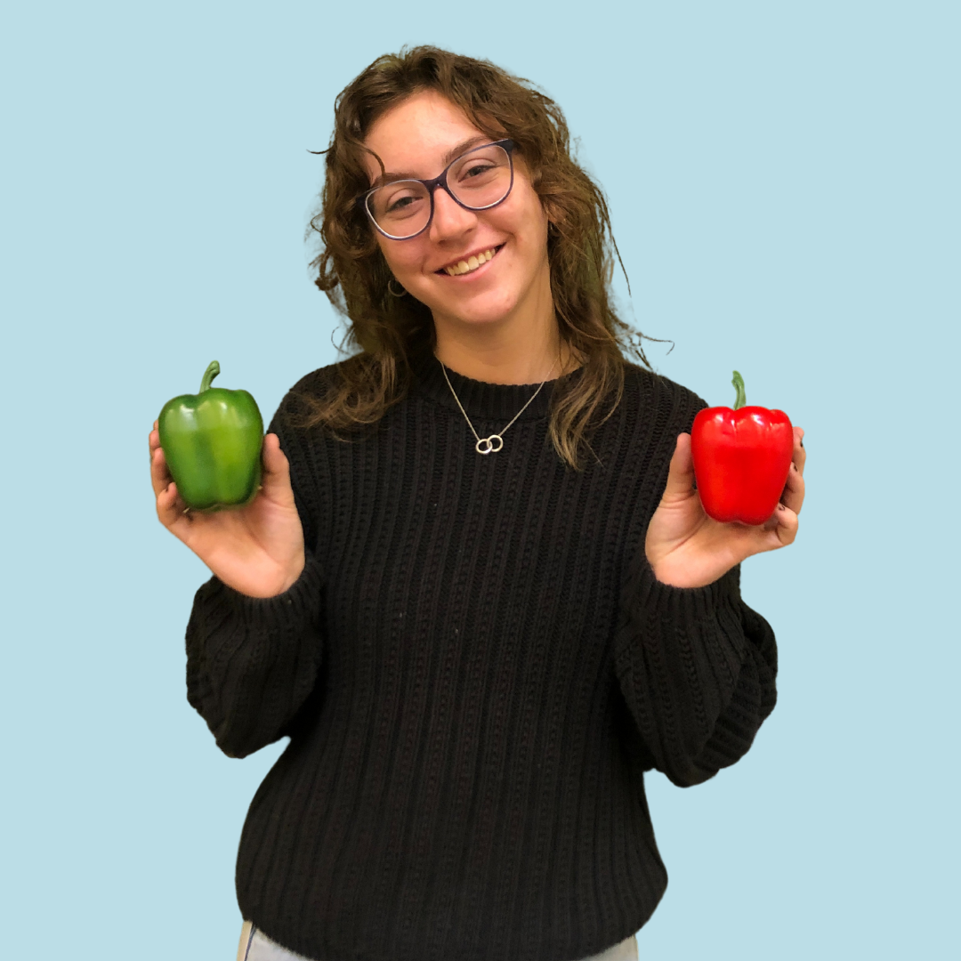 Mary Kate poses holding a green pepper in one hand and red pepper in another. she is wearing a black sweater and has brown wavy hair and wears glasses. 