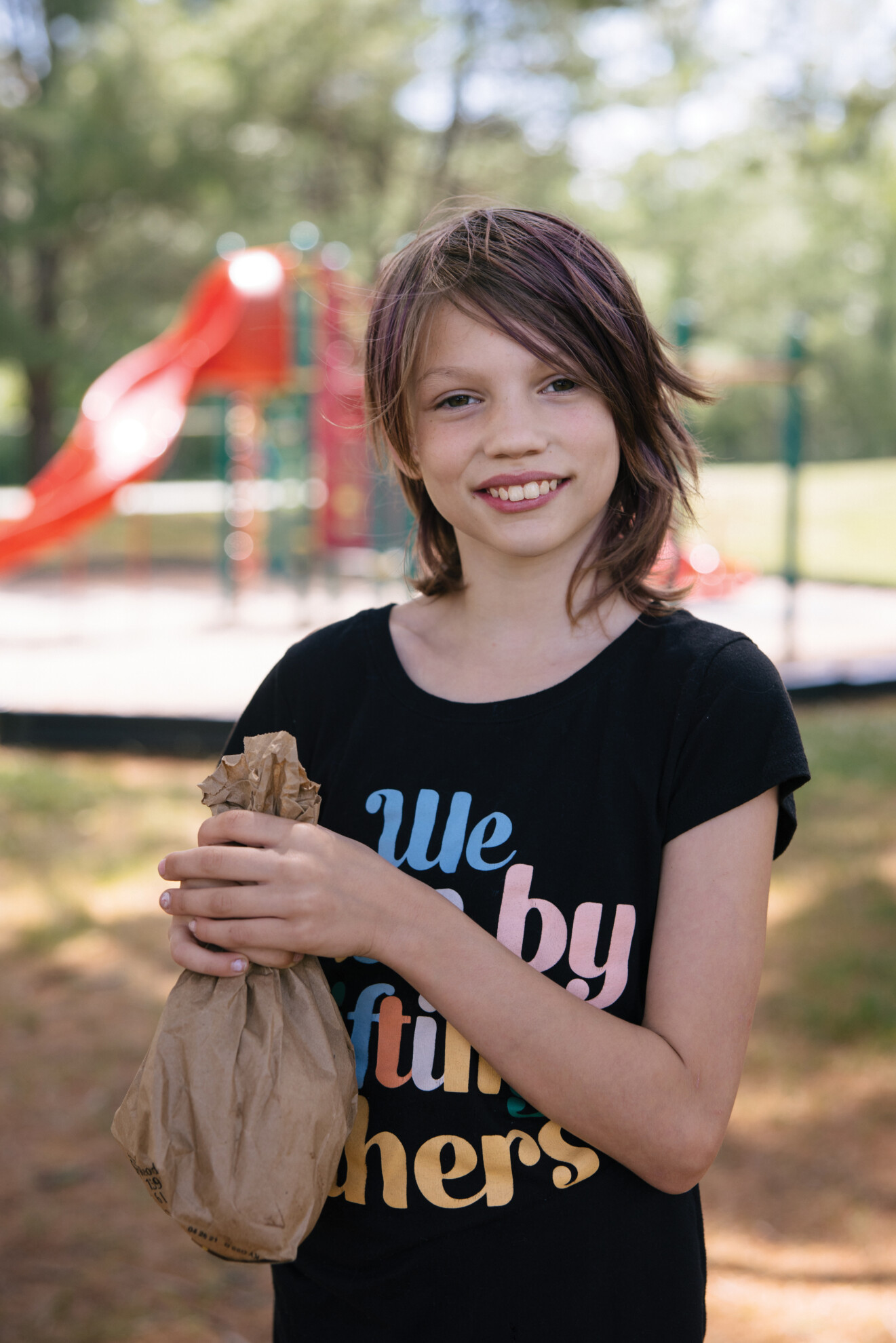 Nine-year-old Marisa stands in front of a park with a brown paper bag lunch. 
