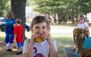 Makaila, a 9 -year-old girl, eats a turkey sandwich in a park with kids playing in the background.
