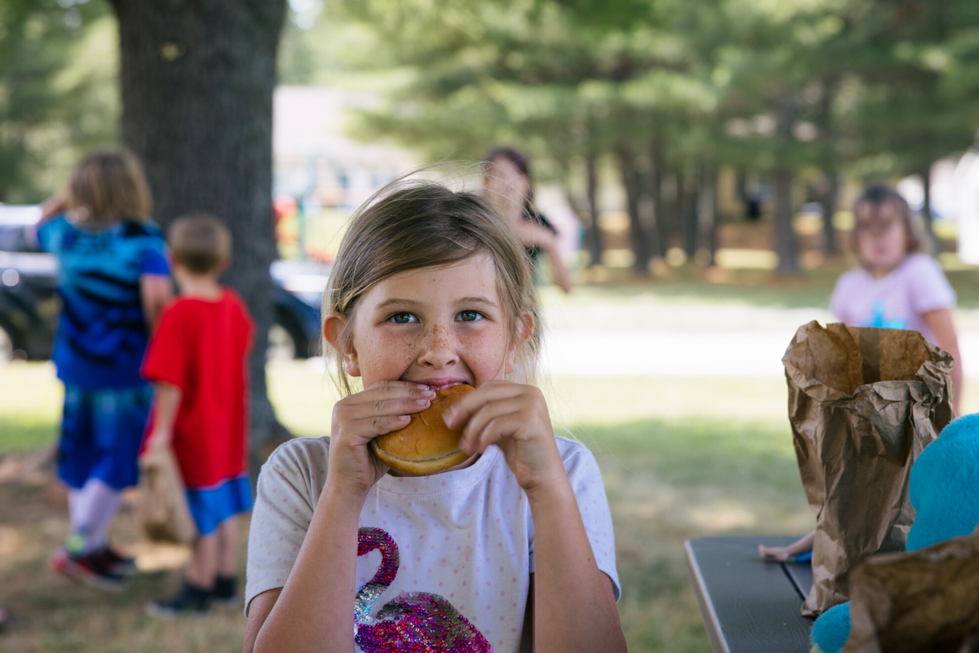 Makaila, a 9 -year-old girl, eats a turkey sandwich in a park with kids playing in the background. 