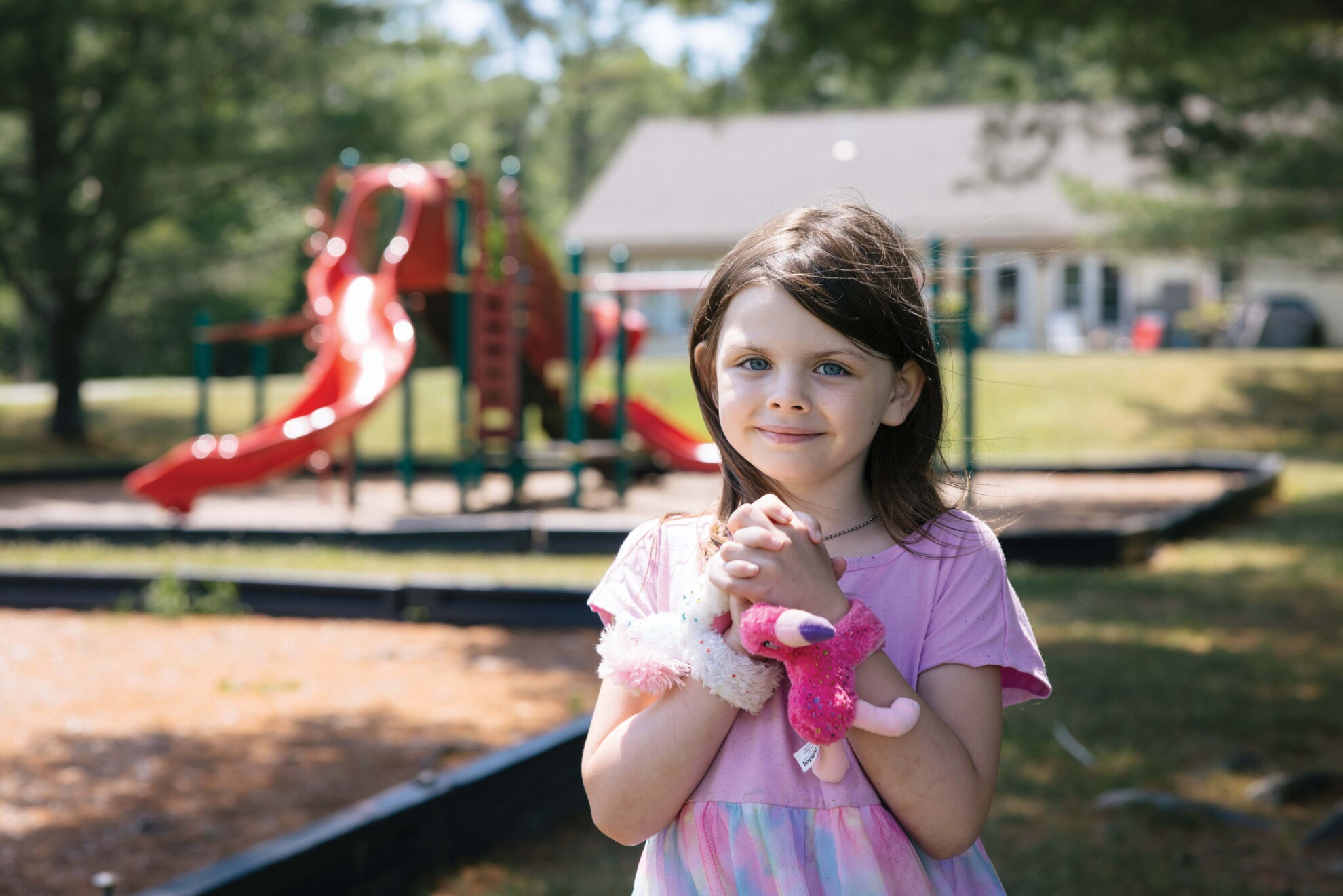 Six-year-old Ami stands in front of a park wearing a pink dress and a flamingo stuffed animal tied around her wrist. 