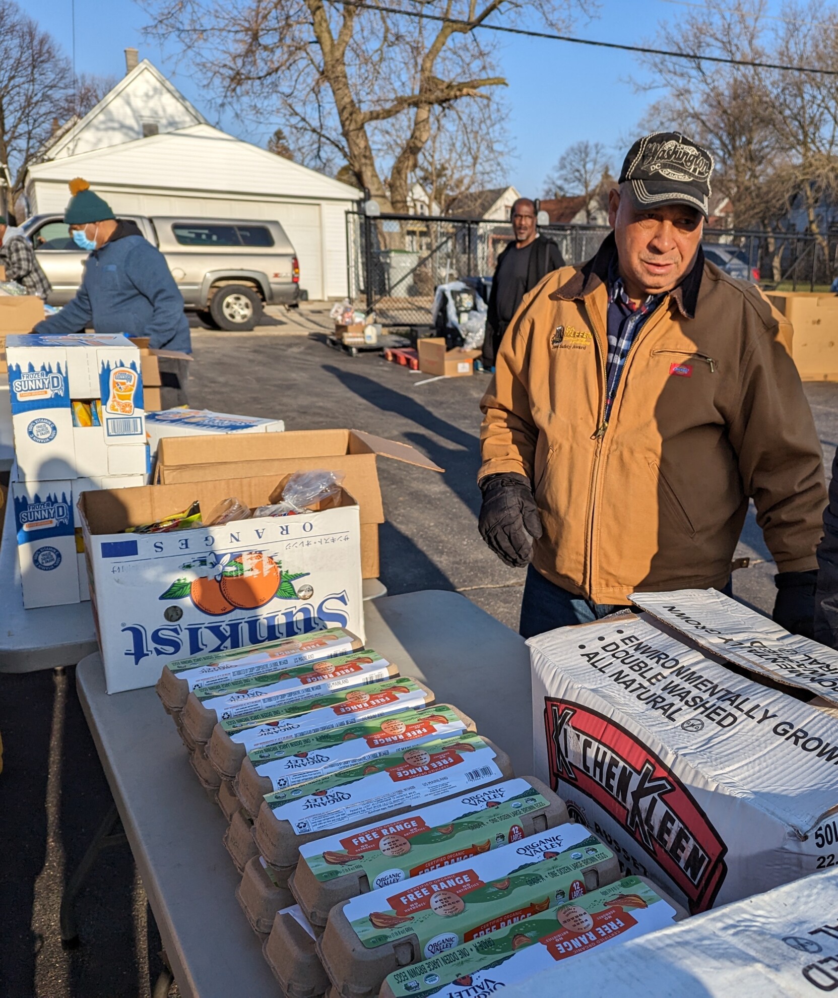 A volunteer wearing a brown jacket and a baseball cap stands by a table filled with eggs ready to distribute them to community members in need.